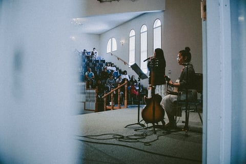 Two people with cello instruments sitting in a bright church interior with tall arched windows and blue decorative panels on the wall