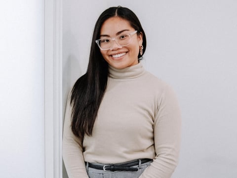 Smiling woman with long black hair, glasses, wearing a cream turtleneck and light blue pants