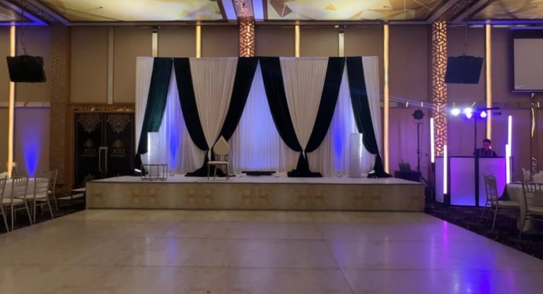 Elegant event venue with stage decorated with blue uplighting, white and black draped curtains, polished floor, and ambient purple lighting