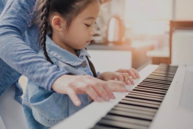 Child learning to play piano with adult guidance, hands positioned on keyboard in bright home setting