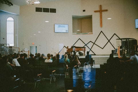Church congregation gathered for worship service with wooden cross, geometric wall art, and seated attendees in rows