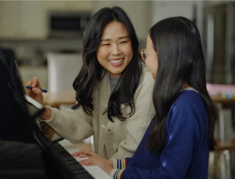 Two young women smiling while playing piano together in a bright room