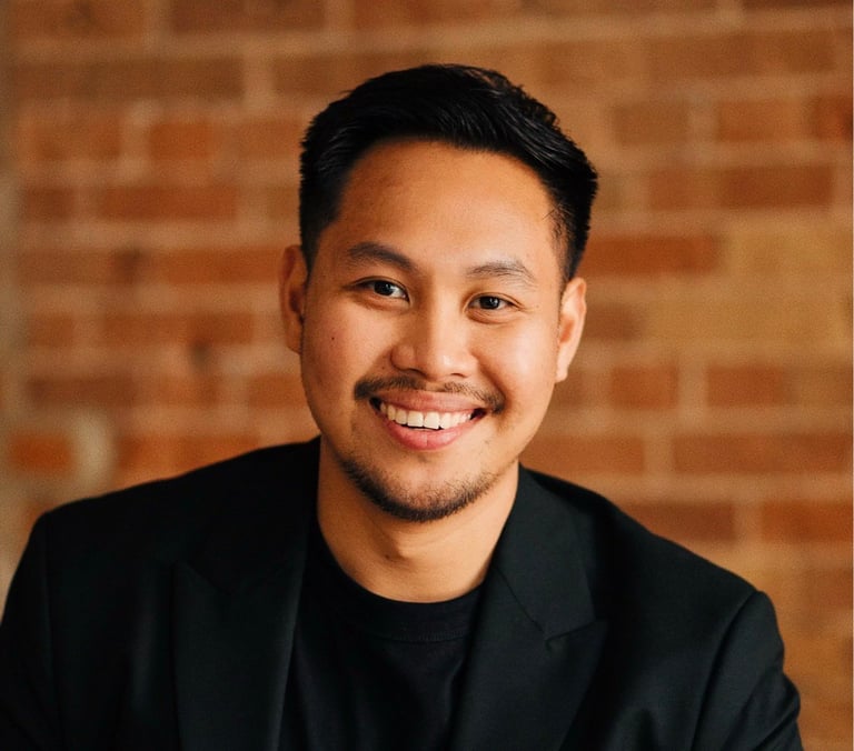 Man with black hair smiling at the camera wearing a black blazer over a black shirt against a brick wall background